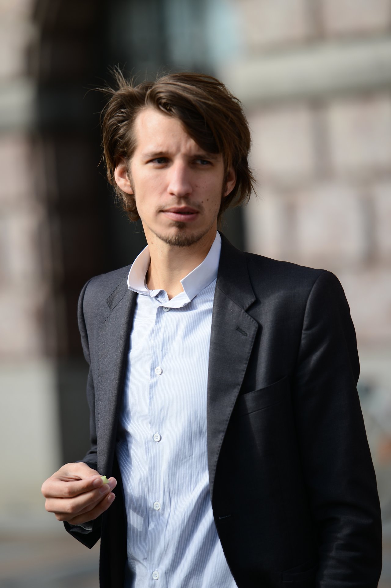 A man in a suit stands outside, looking to the side while holding a small object in his hand.