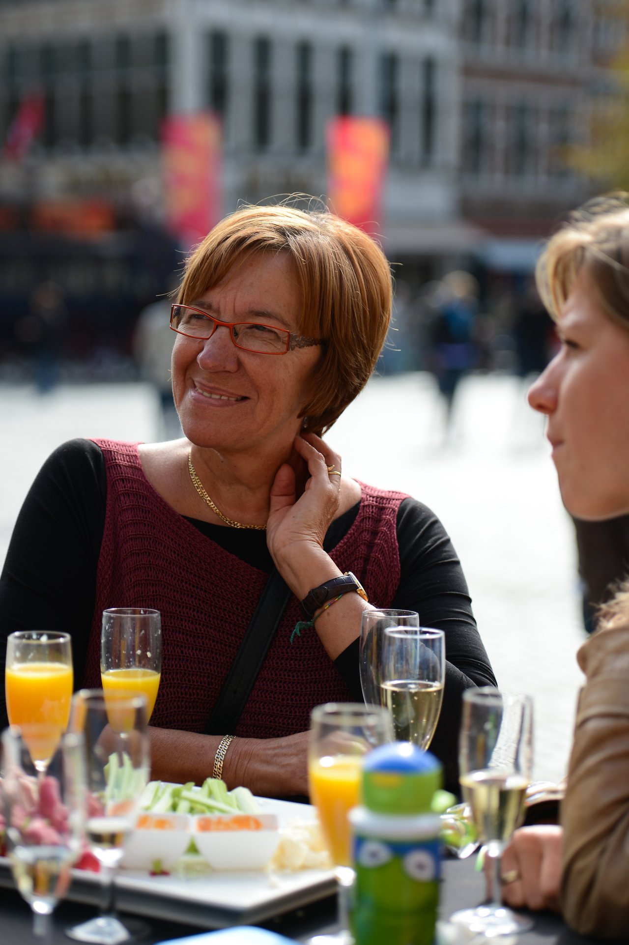 A woman wearing glasses smiles while sitting at an outdoor table with drinks and food, talking to another person.