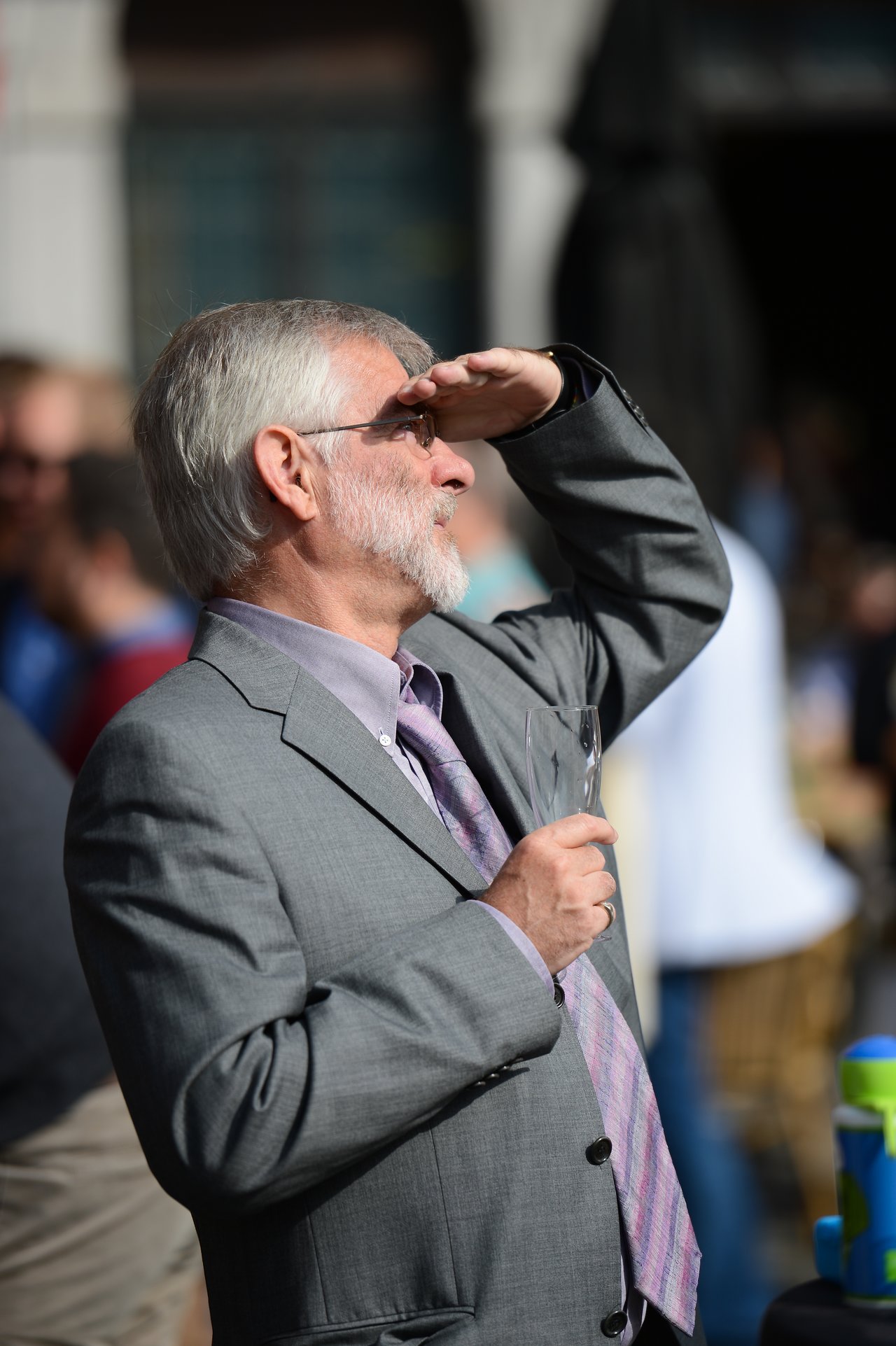 An older man in a suit holds a champagne glass and shades his eyes while looking into the distance.