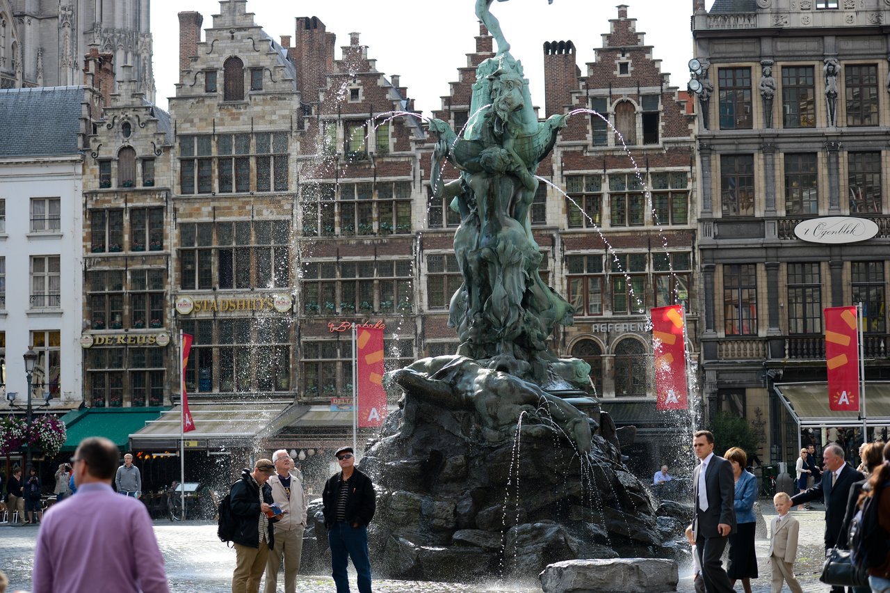 A fountain with a statue in a busy city square, surrounded by people walking and talking.