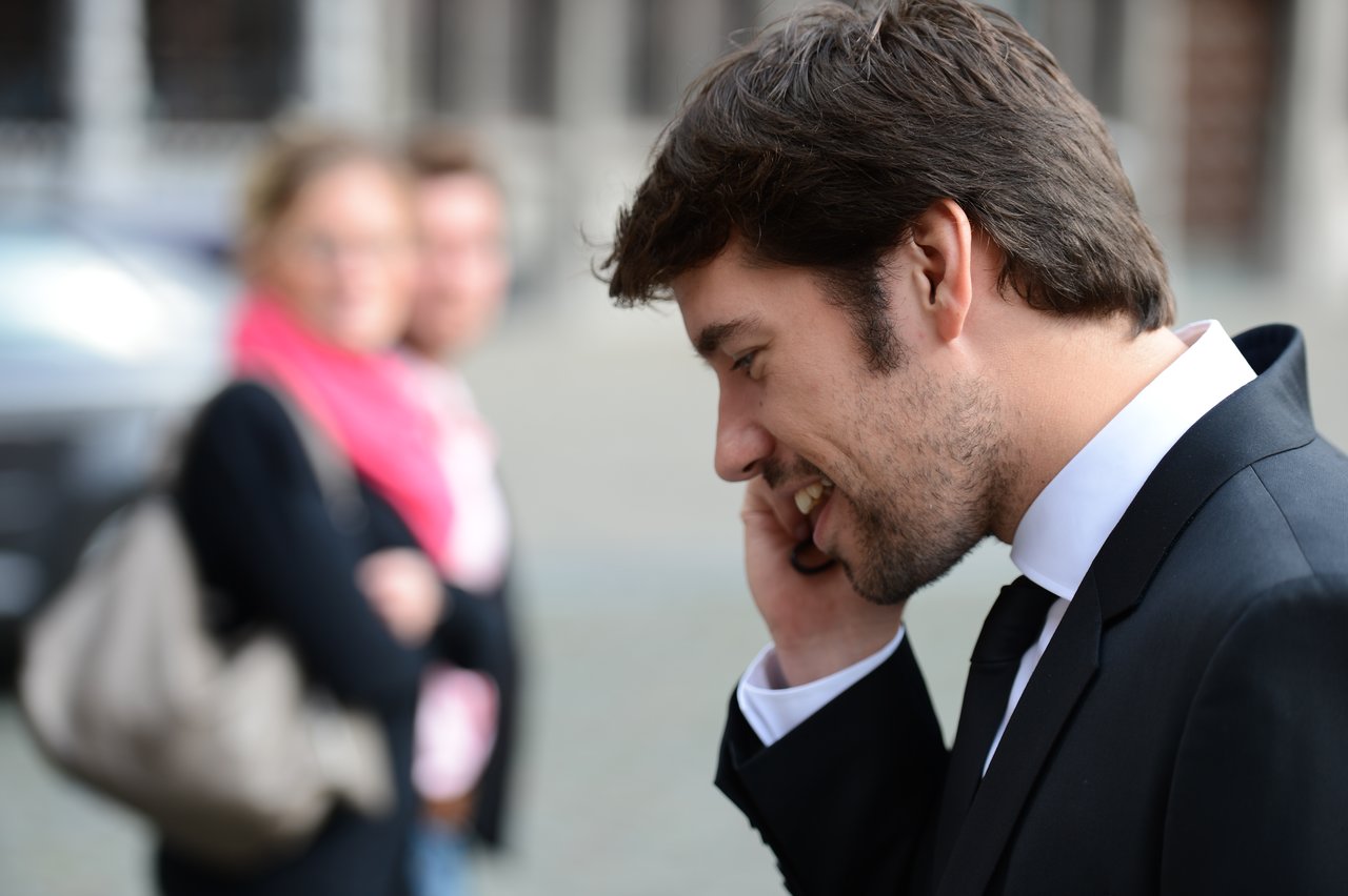 A man in a suit smiles while talking on the phone, with two blurred people in the background.