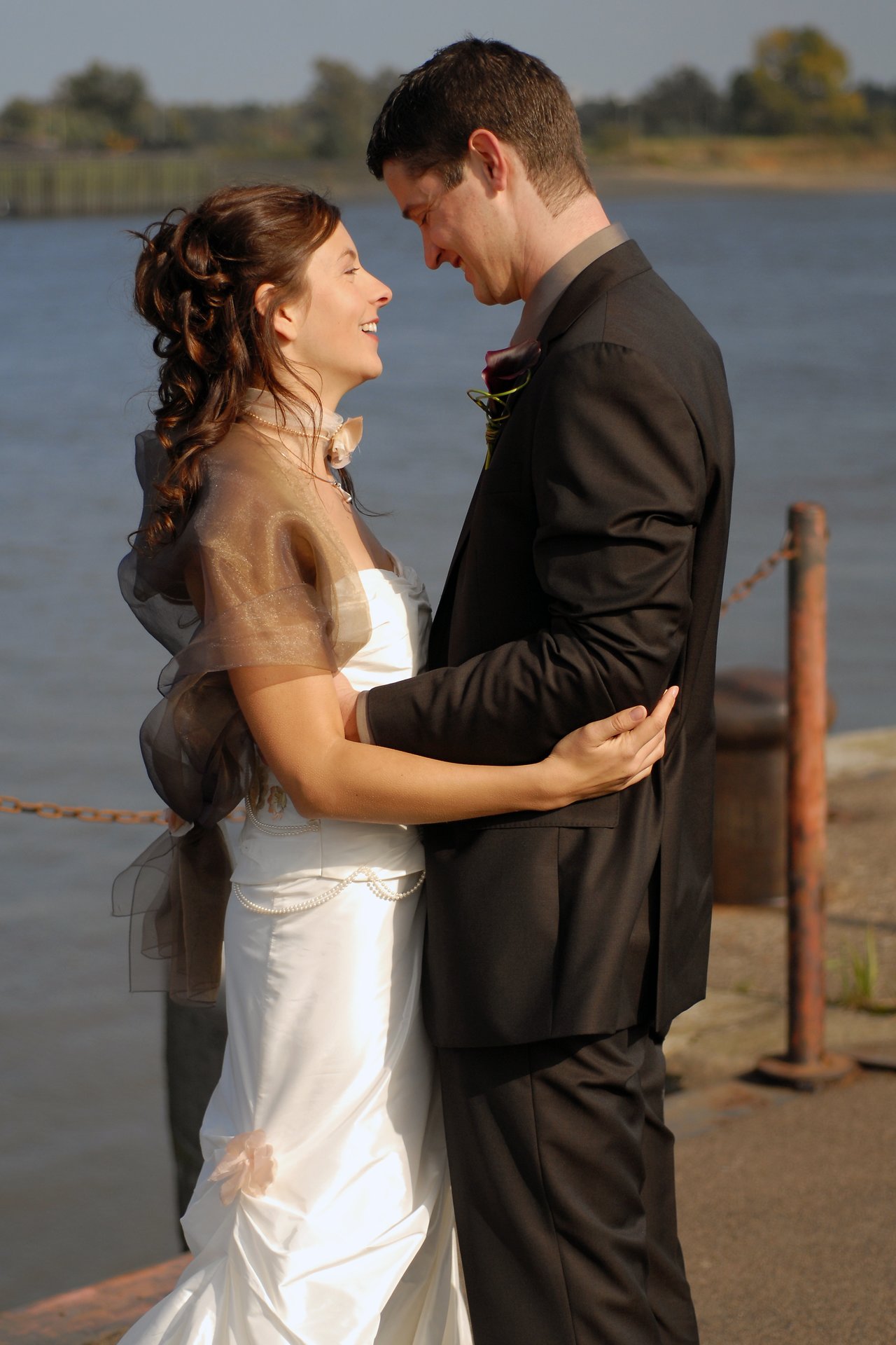 A bride and groom stand close together, smiling at each other on their wedding day near the water.