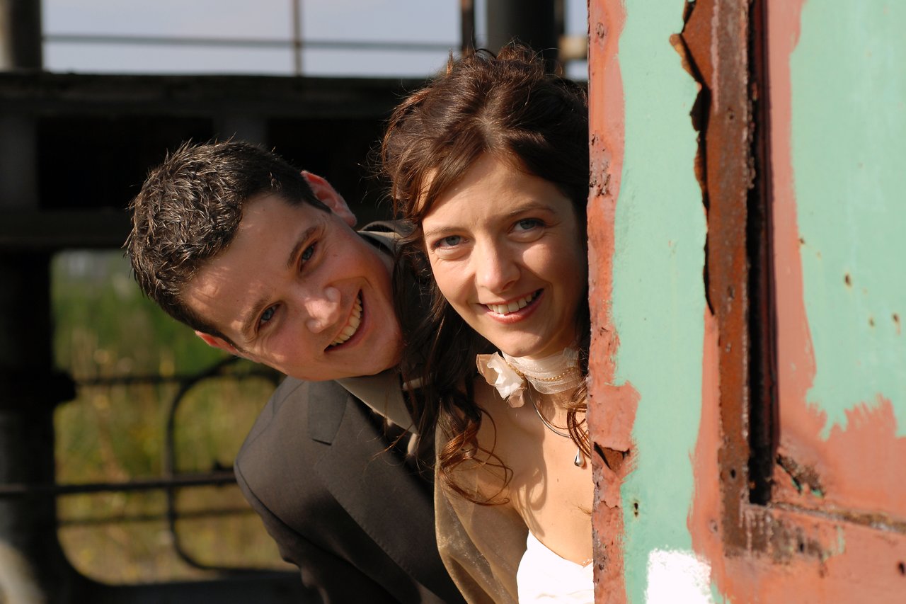 A smiling bride and groom peek out from behind a weathered metal structure on their wedding day.