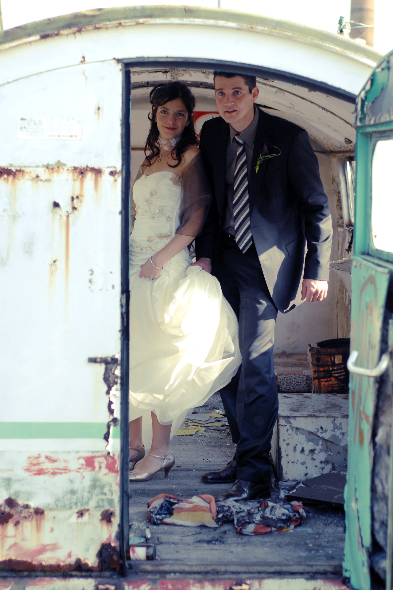 A bride and groom stand together inside an old, rusted vehicle, smiling at the camera.