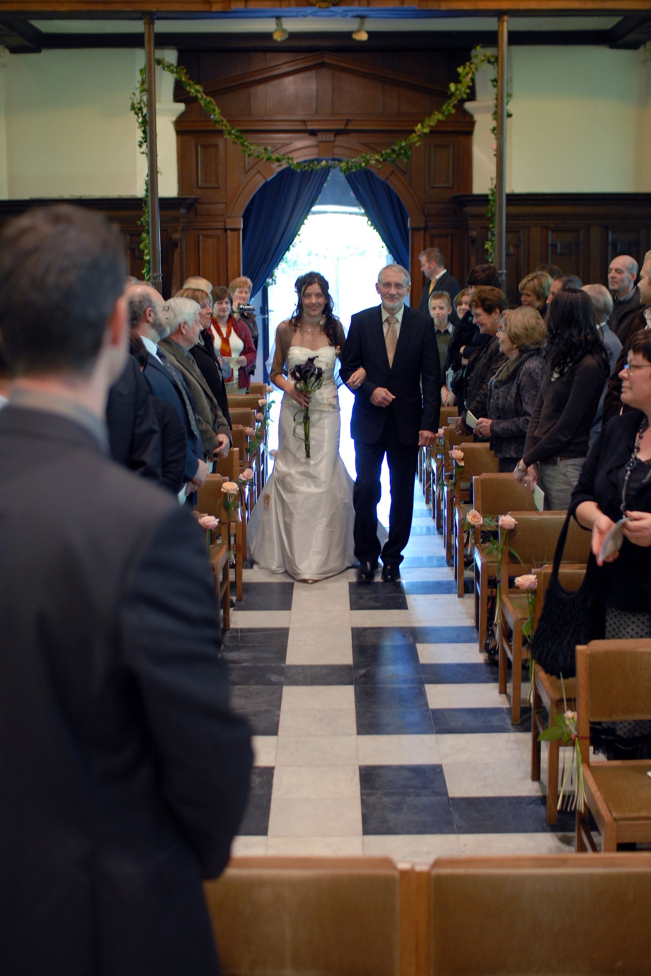 A bride in a white dress walks down the aisle with an older man, surrounded by seated wedding guests.
