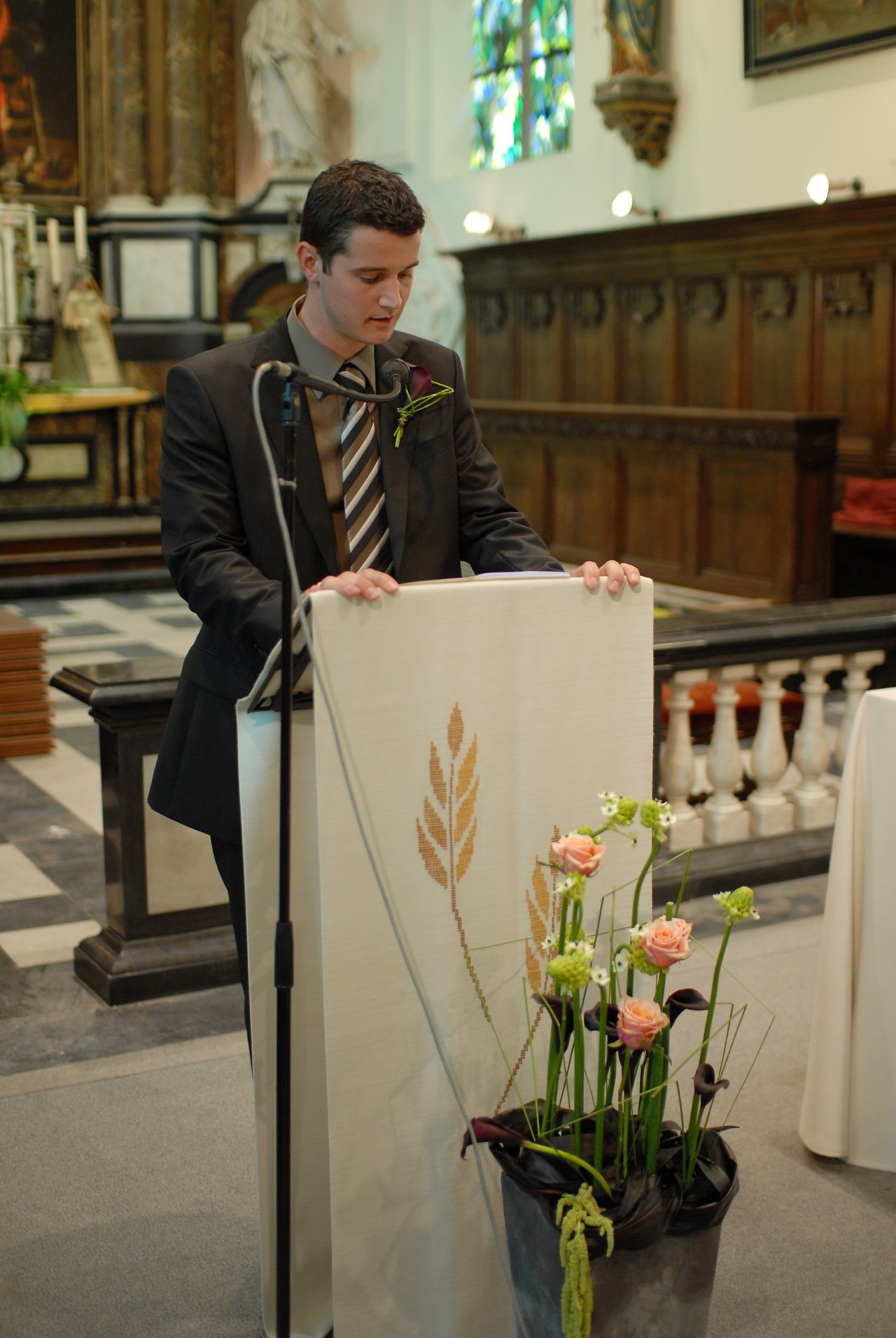 A man in a suit stands at a lectern in a church, reading or speaking into a microphone.