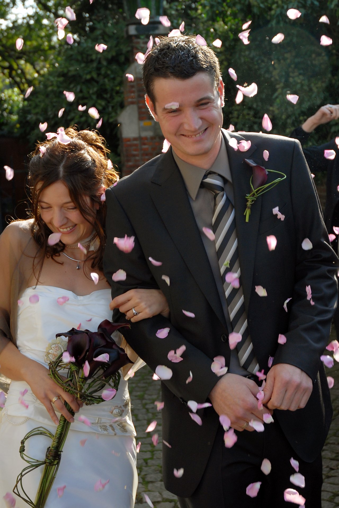 A smiling bride and groom walk arm in arm as pink flower petals are thrown around them.