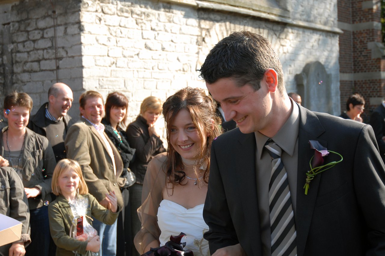 A smiling bride and groom walk together outside, surrounded by friends and family celebrating their wedding.