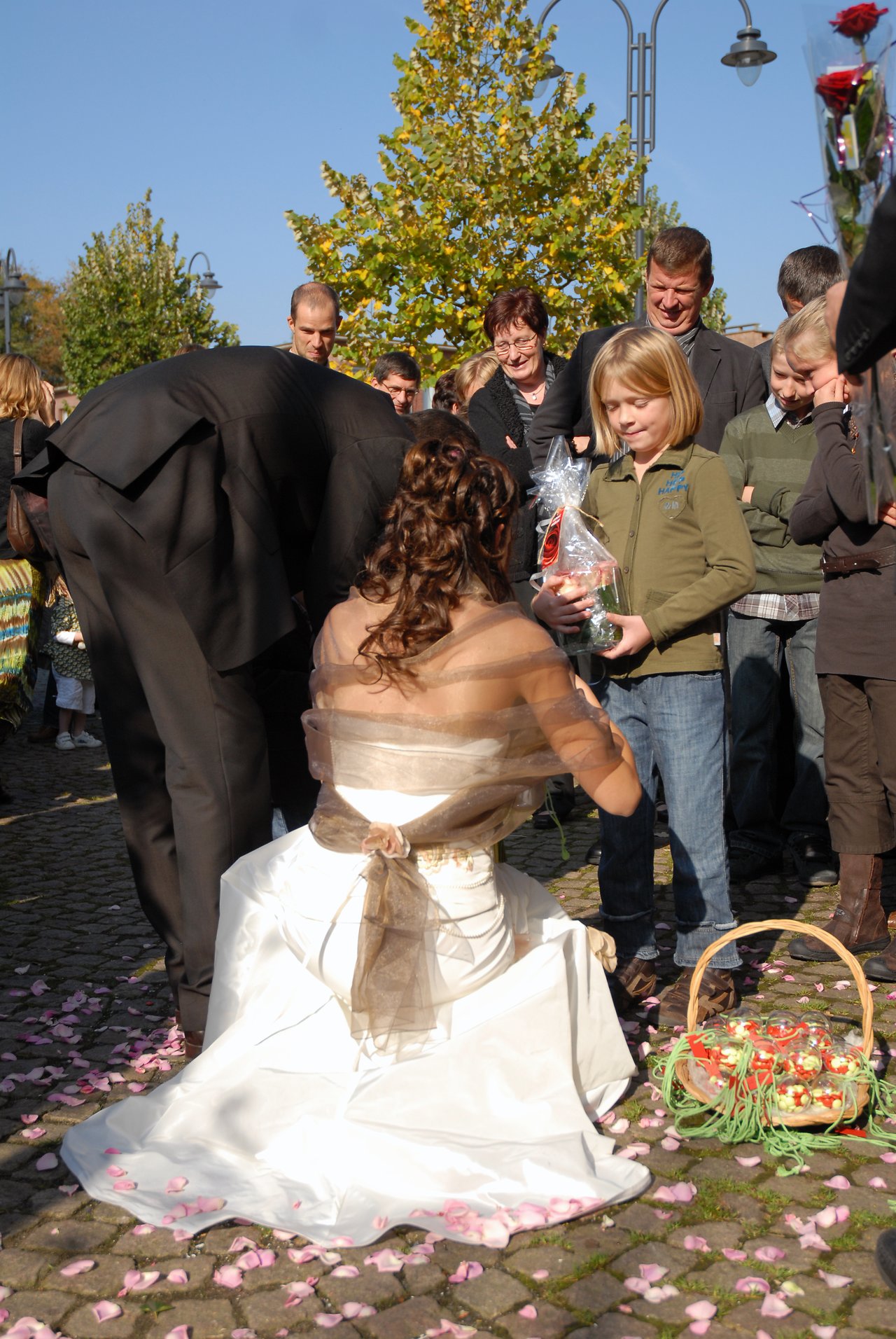 The bride kneels on the ground, receiving a gift from a young girl, surrounded by wedding guests.