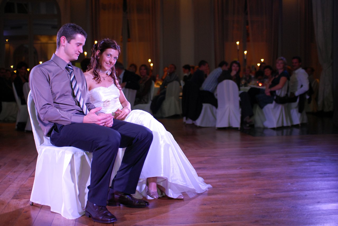 A bride and groom sit together on a dance floor, smiling, while wedding guests watch from their tables.