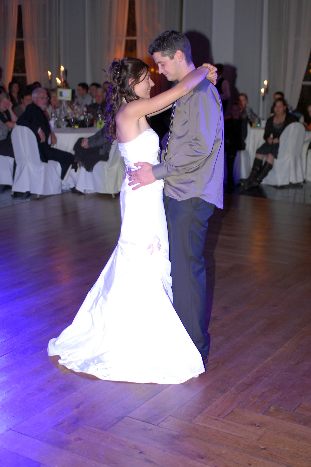 The bride and groom share their first dance, embracing and smiling, while wedding guests watch from their tables.