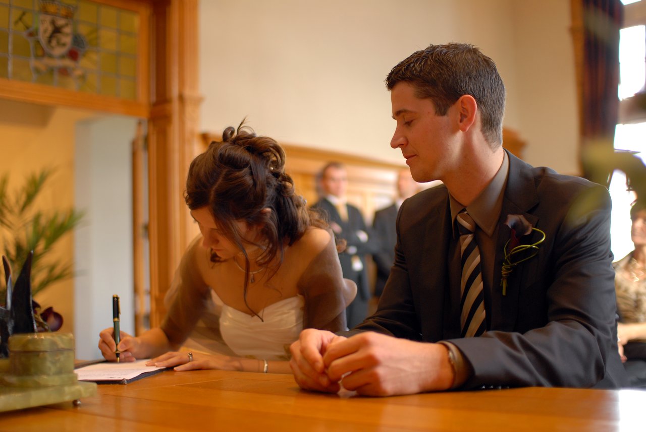 The bride signs a document while the groom sits beside her at a wedding ceremony.