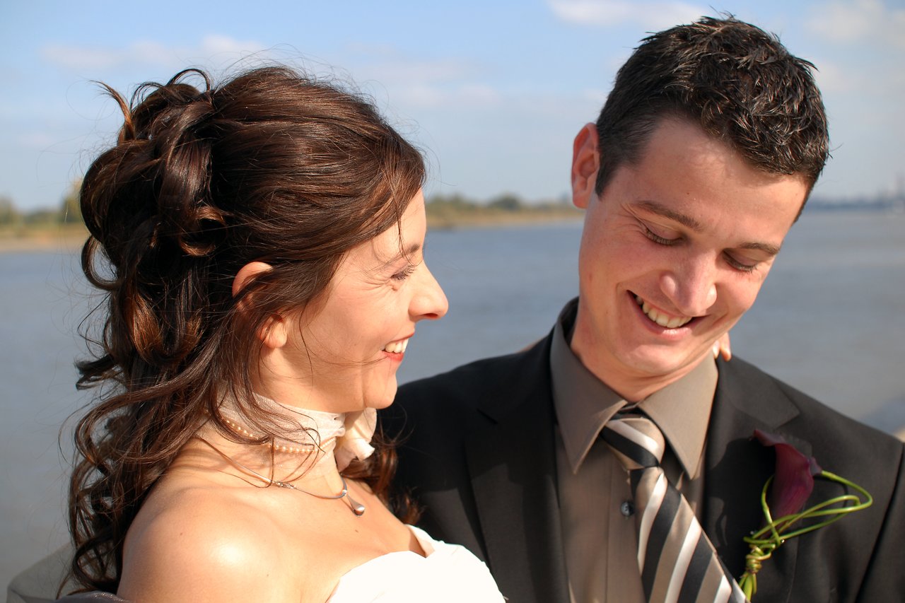A bride and groom smile at each other outdoors, with the groom looking down and the bride leaning toward him.