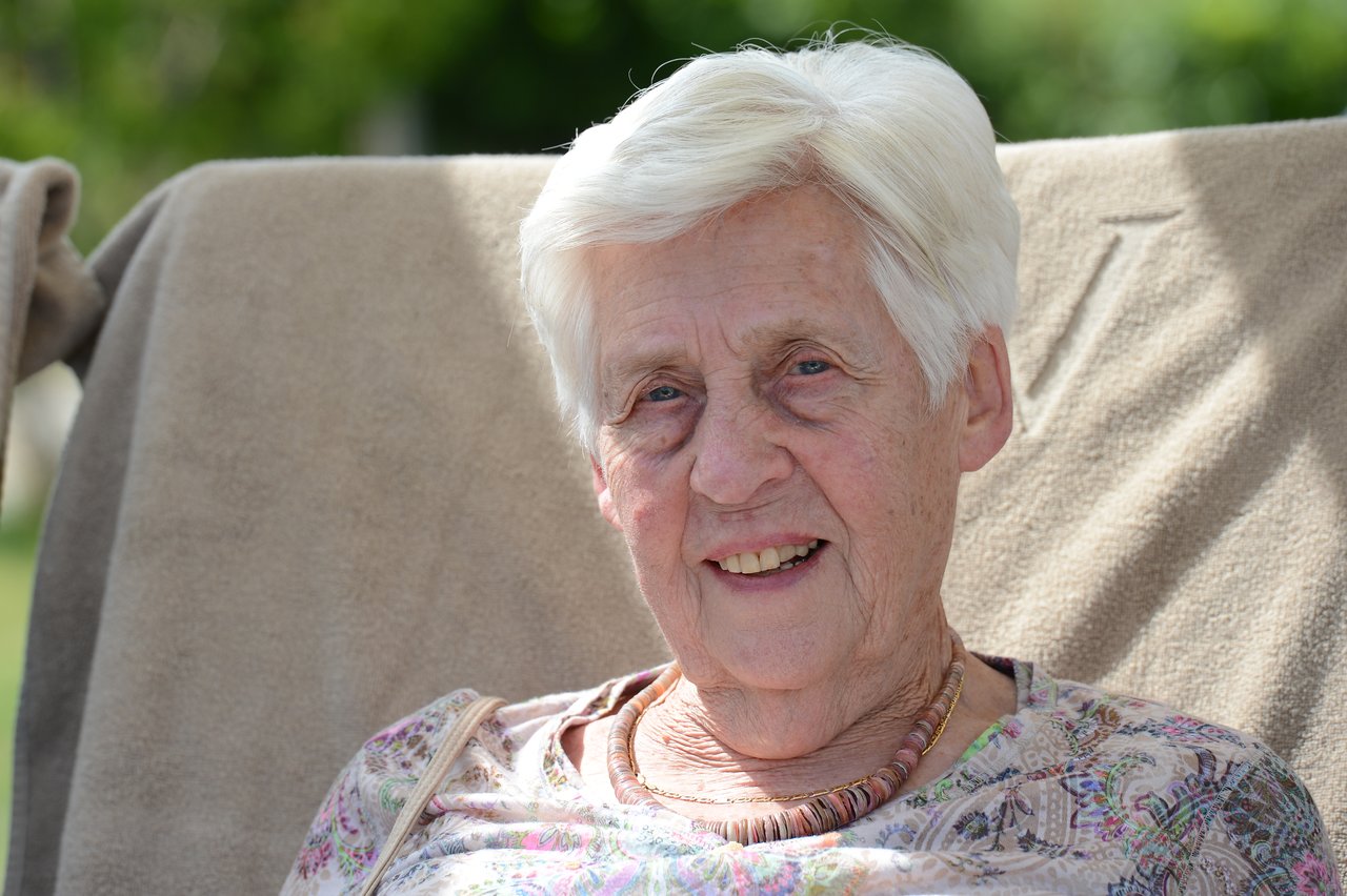 Elderly woman with white hair sits outdoors on a chair, smiling at the camera.