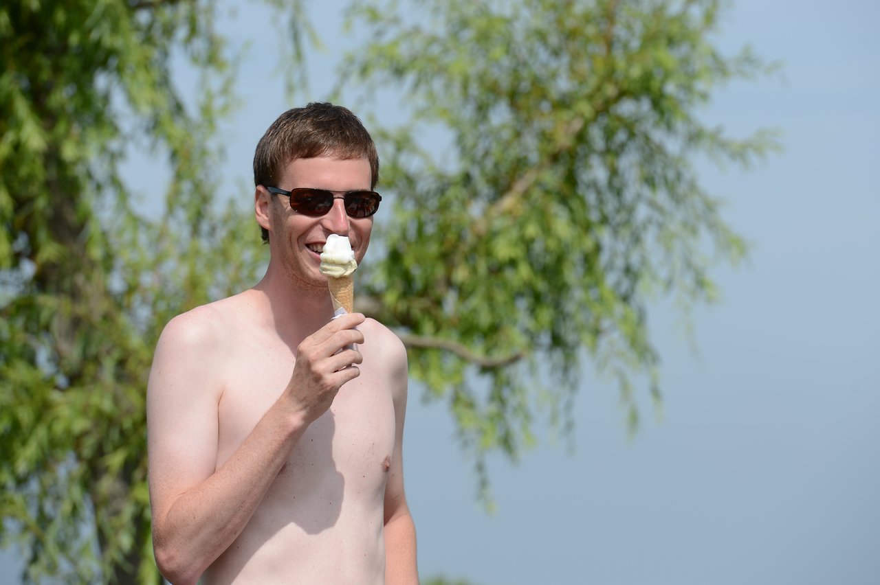A shirtless man wearing sunglasses smiles while holding an ice cream cone outdoors.