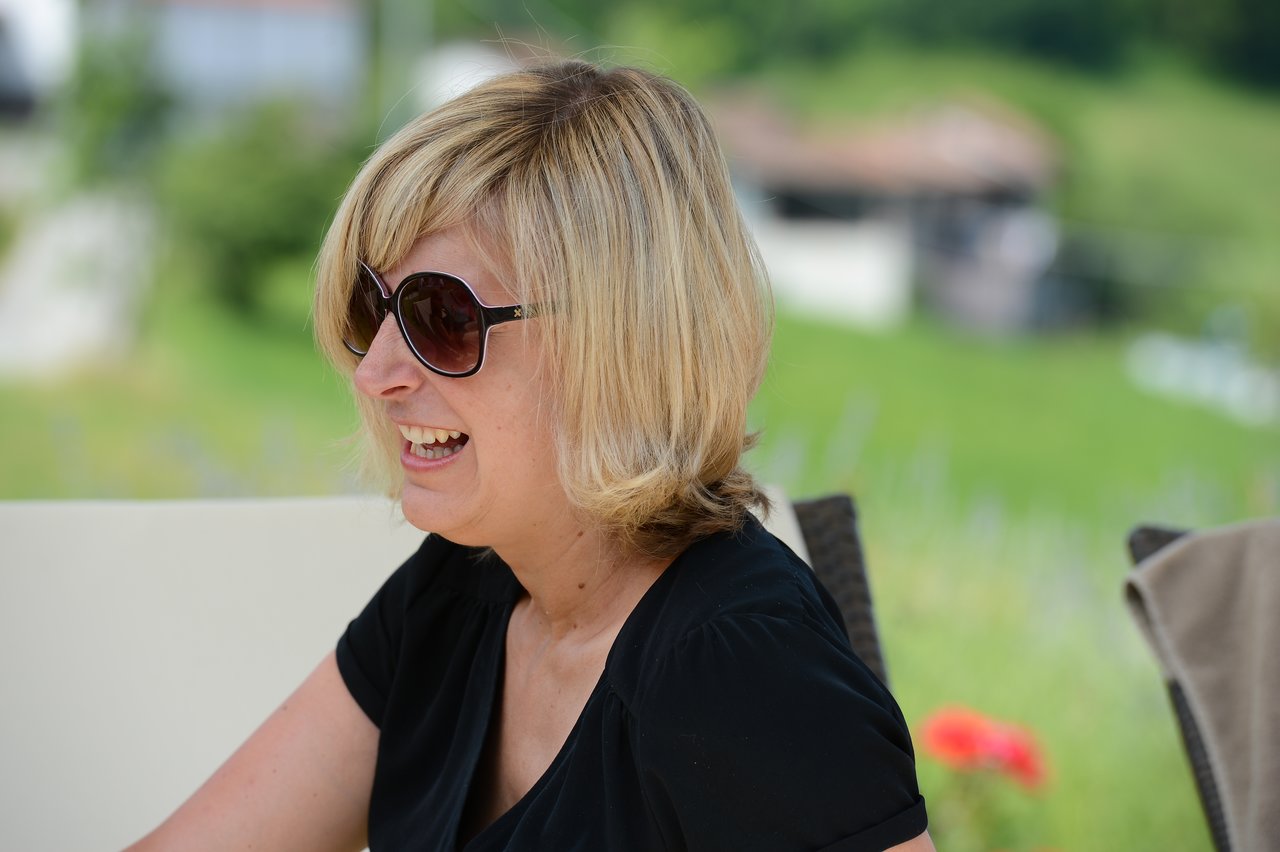 A woman wearing sunglasses and a black dress smiles while sitting outdoors at a wedding.