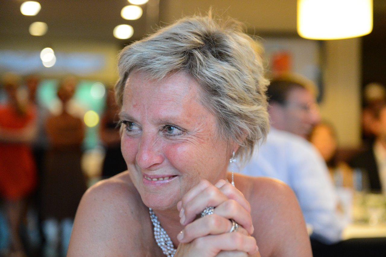 A woman with short blonde hair smiles and clasps her hands at a wedding reception.