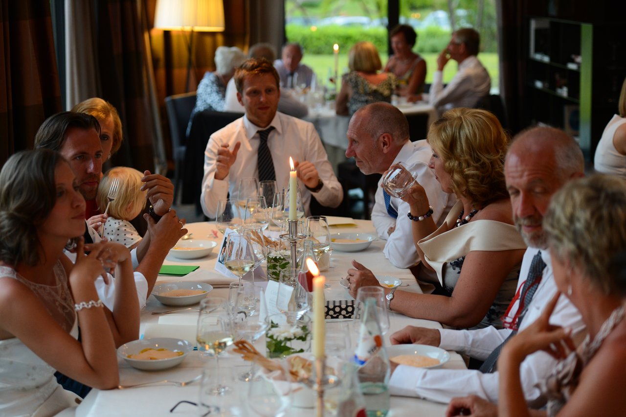 Wedding guests sit at a decorated table, eating, drinking, and talking during a reception dinner.