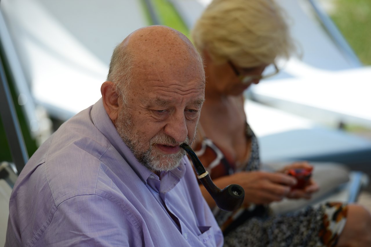 An older man in a light purple shirt smokes a pipe, while a woman behind him looks at her phone.