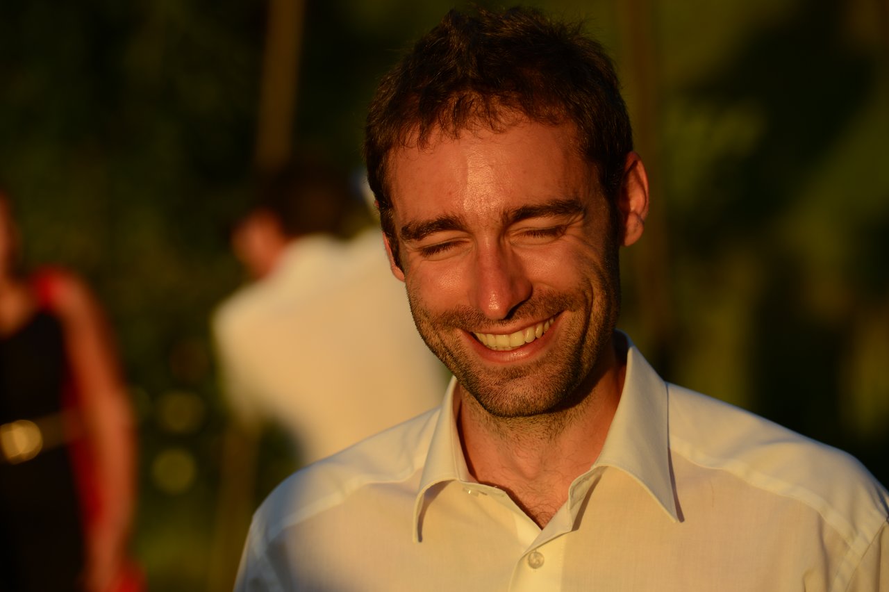 A man in a white shirt smiles with his eyes closed, standing outdoors in warm sunlight.
