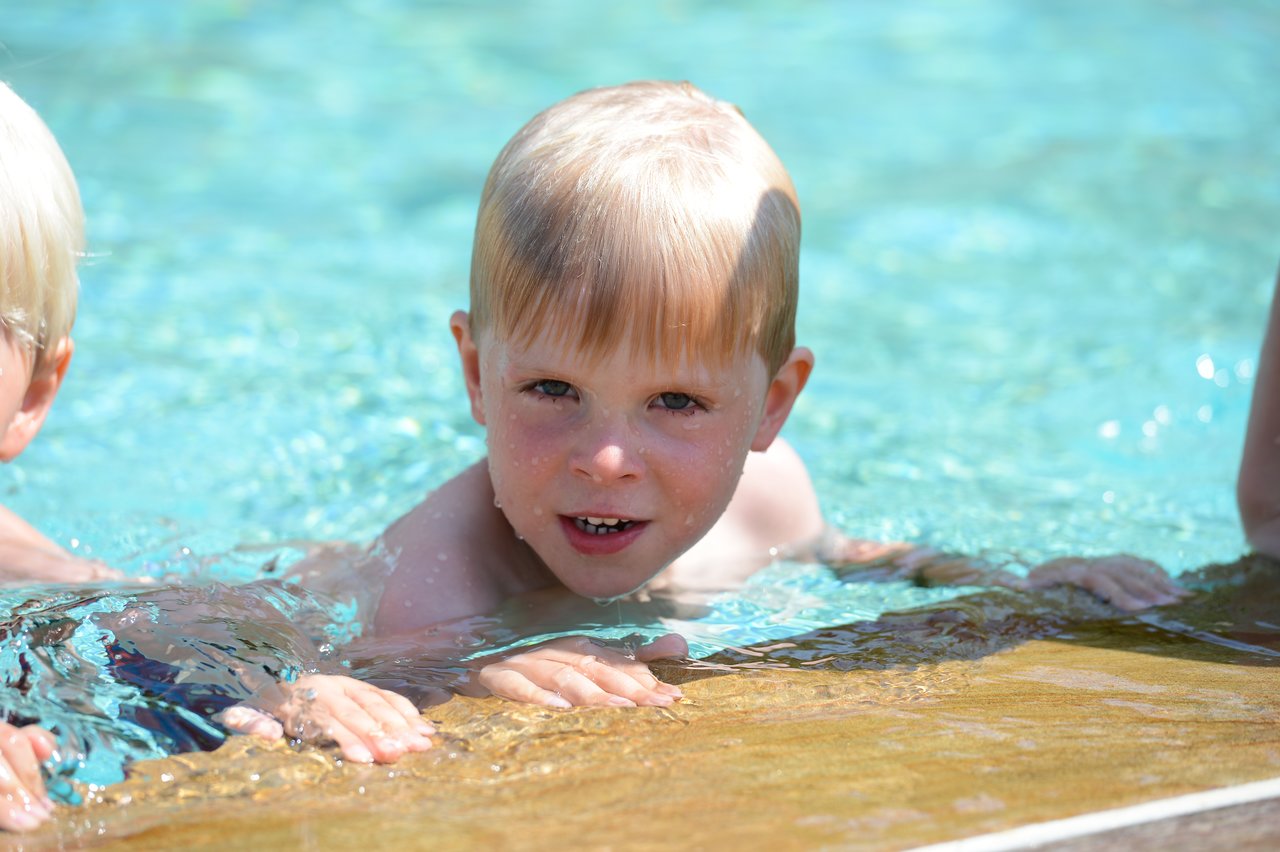 A young child with wet hair holds onto the pool edge, looking forward while swimming.