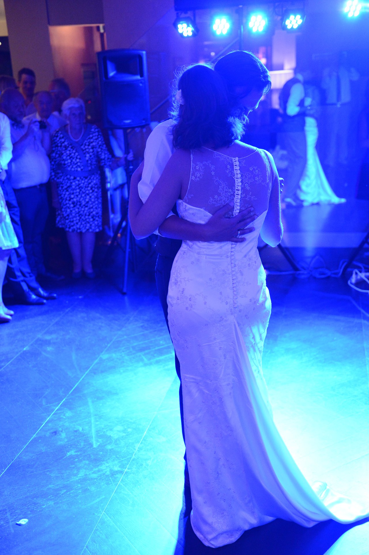 The bride and groom share their first dance, embracing on the dance floor under blue lighting, with guests watching.