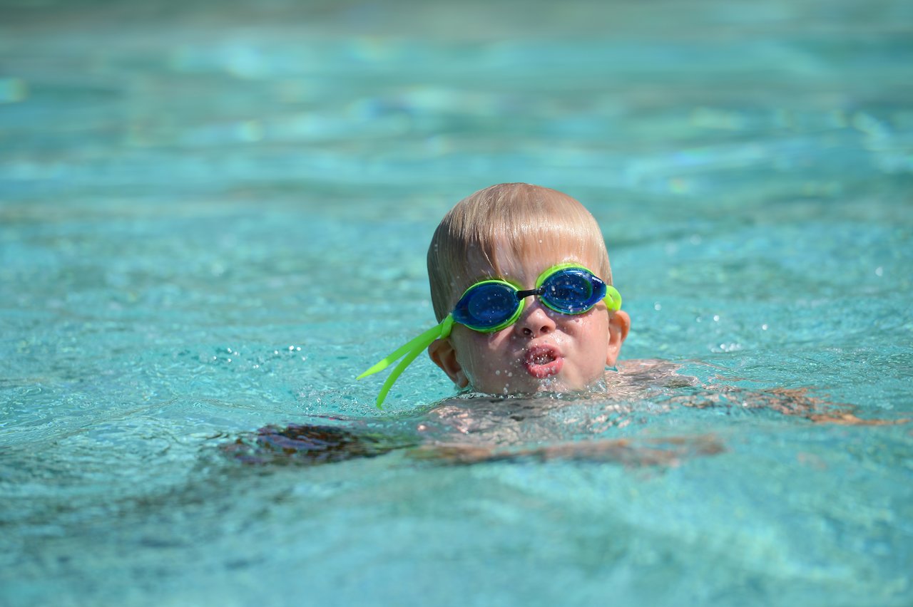 A young child wearing goggles swims in a pool, with their face partially above the water.