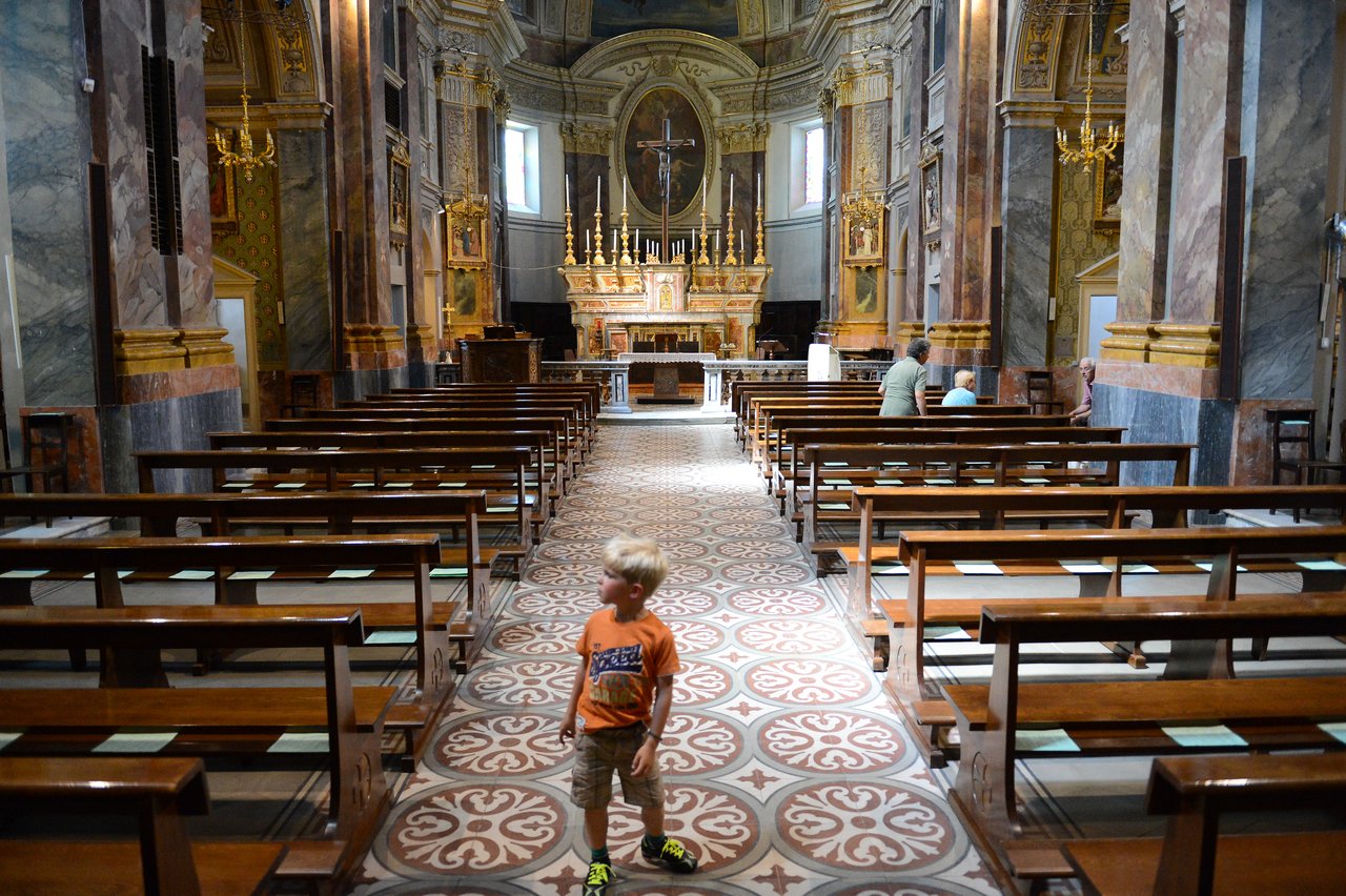 A young child walks down the aisle of a church, while a few people sit on the benches.