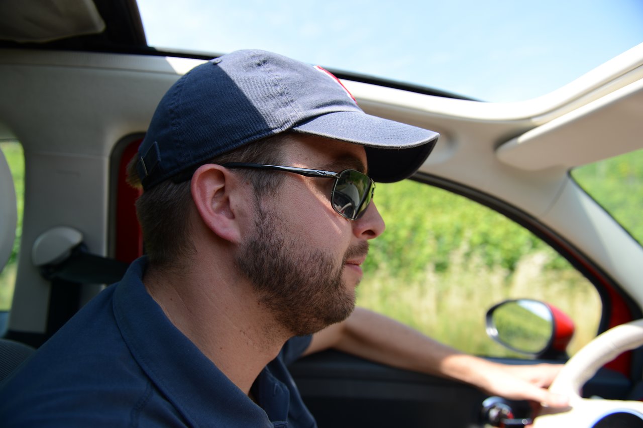 A man wearing sunglasses and a cap is driving a car with an open sunroof.