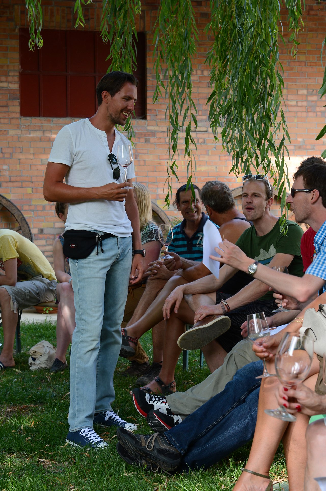 A man in a white shirt stands holding a wine glass, smiling and talking with seated guests at a wedding.