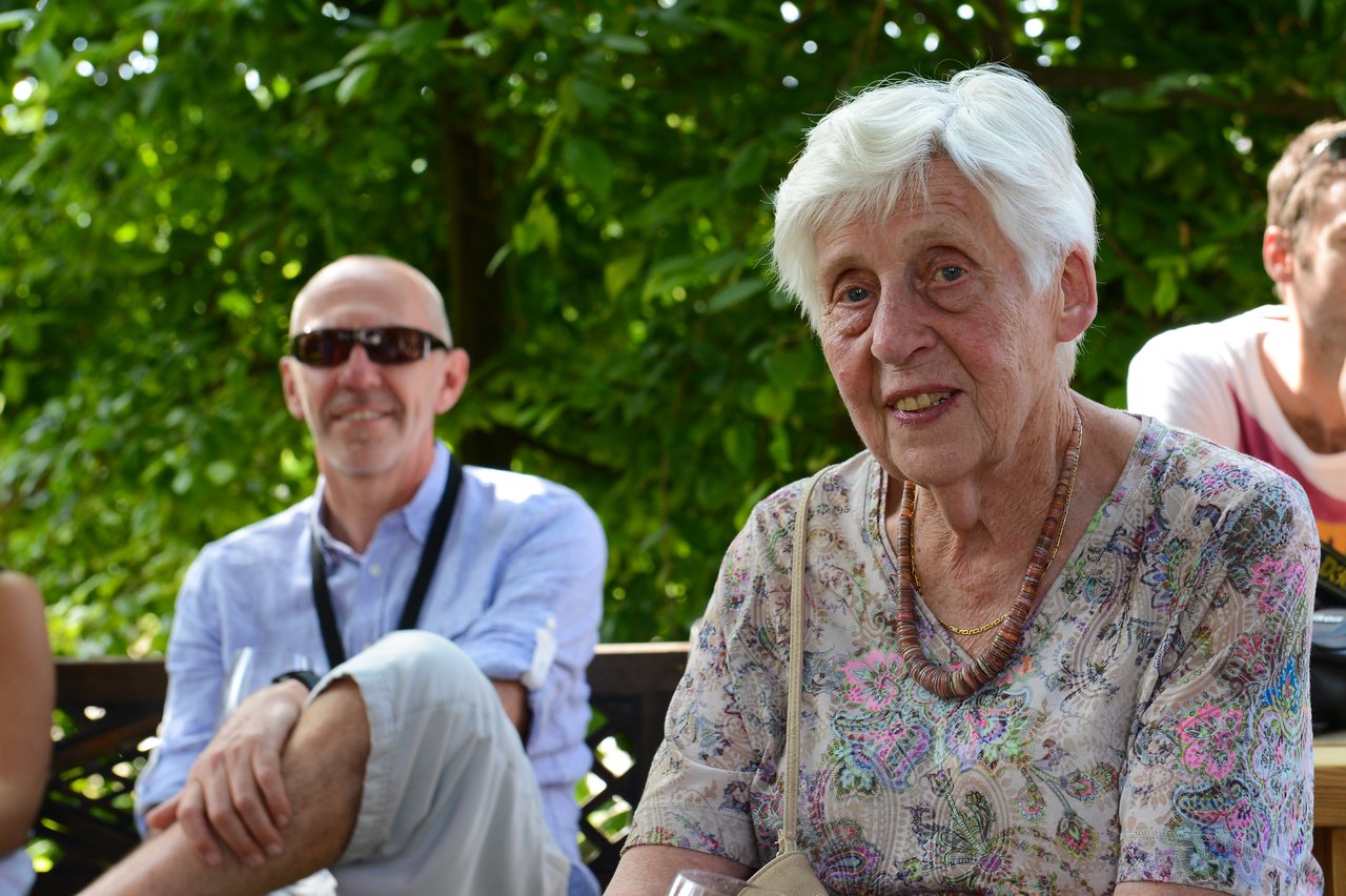 An elderly woman smiles at the camera while seated outdoors, with a man in sunglasses sitting behind her.