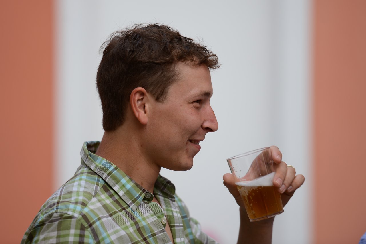 A man in a plaid shirt holds a glass of beer and smiles while looking to the side.