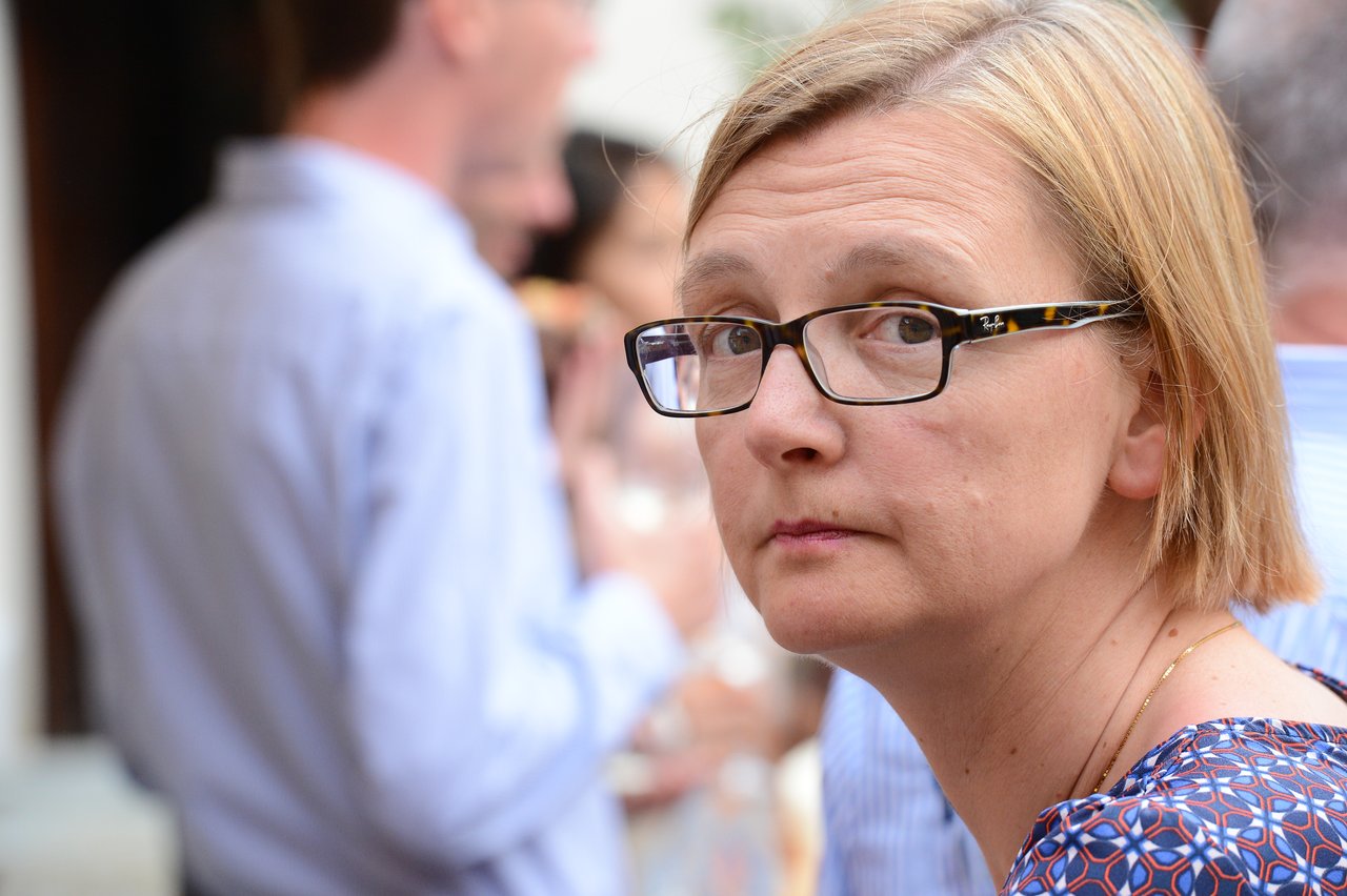 A woman with glasses looks toward the camera at a wedding reception, with guests socializing in the background.