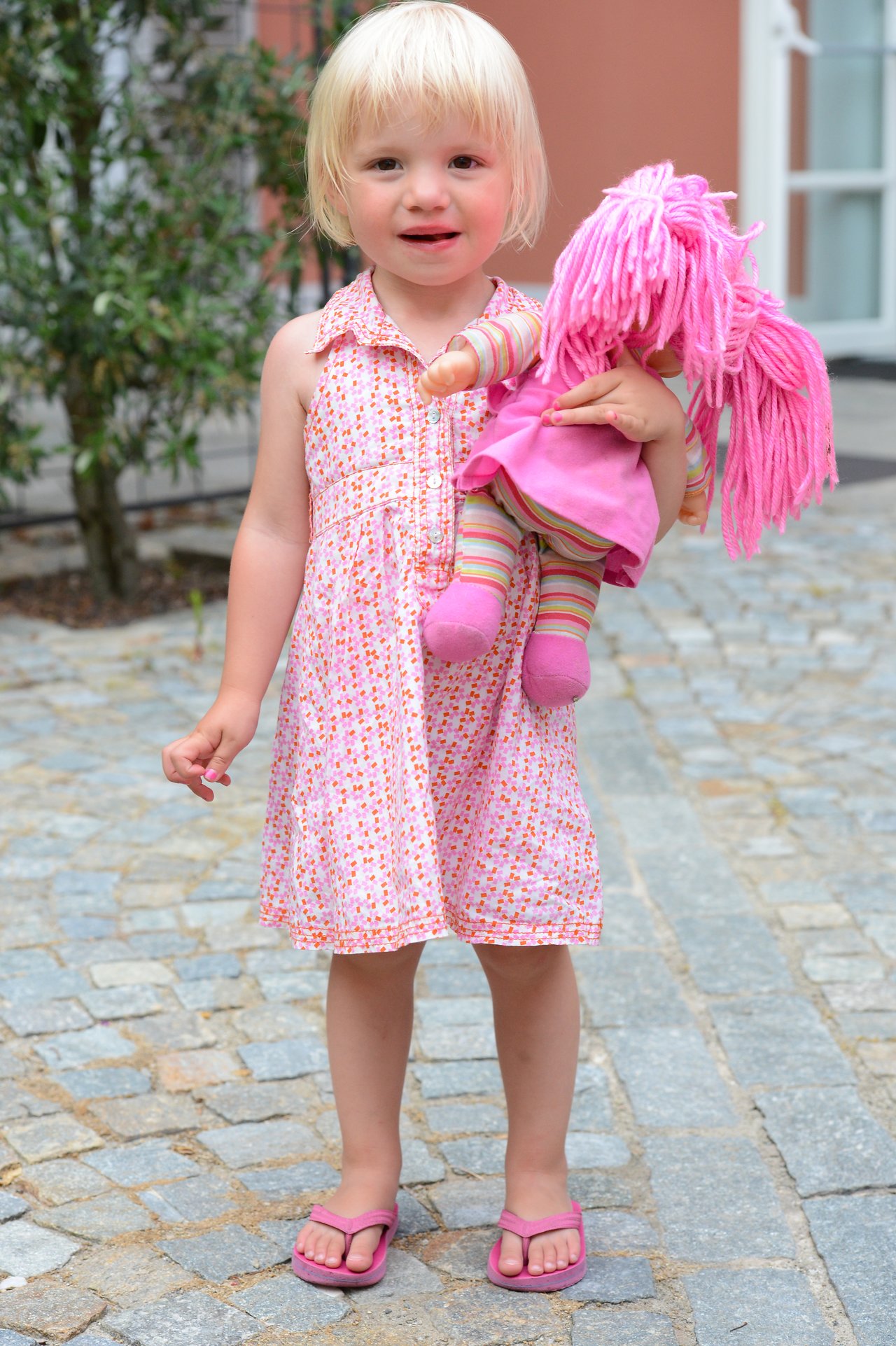 A young child in a pink dress holds a doll with pink yarn hair, standing on a cobblestone path.