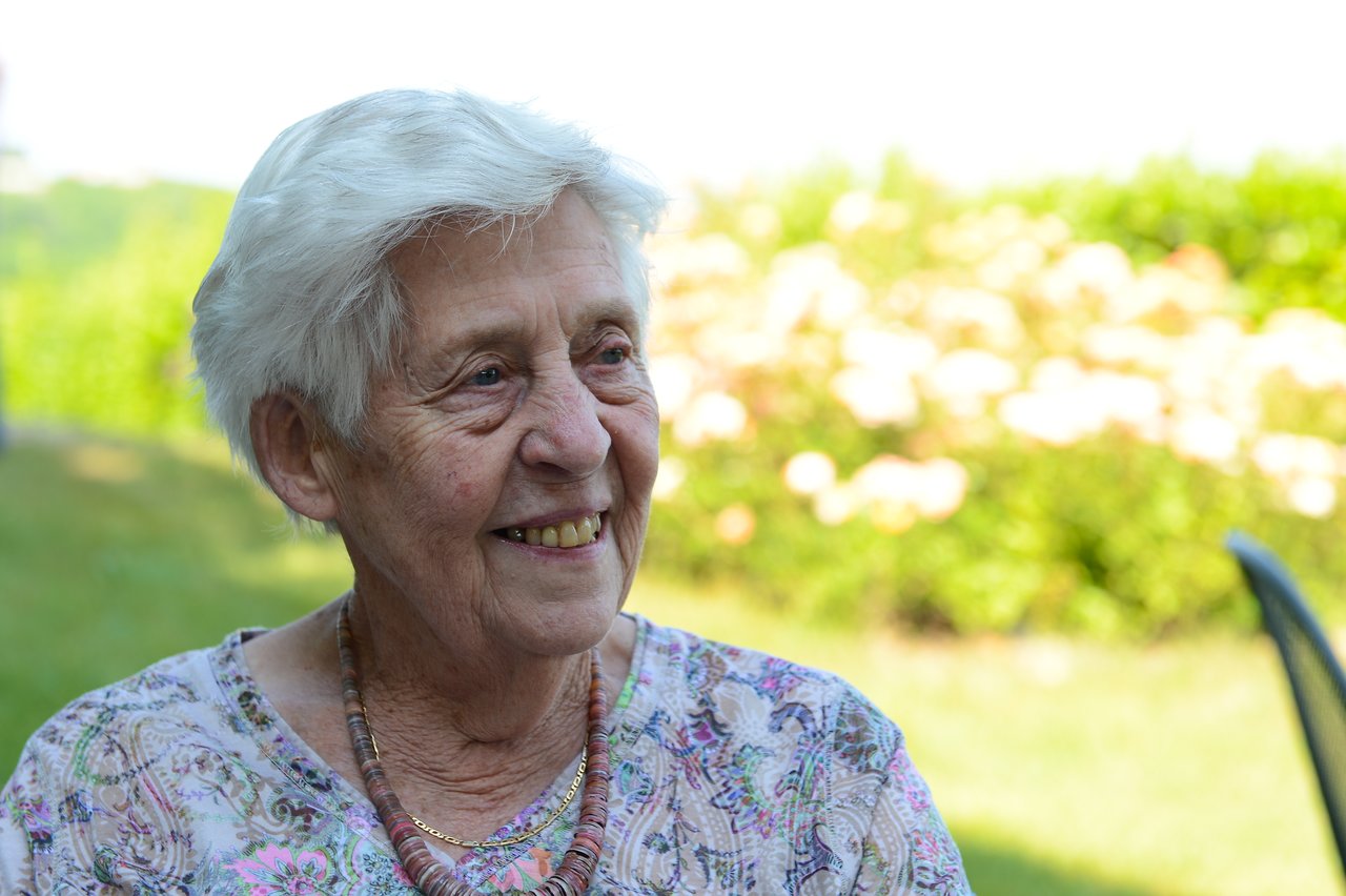 An elderly woman with short white hair smiles while sitting outdoors at a wedding.