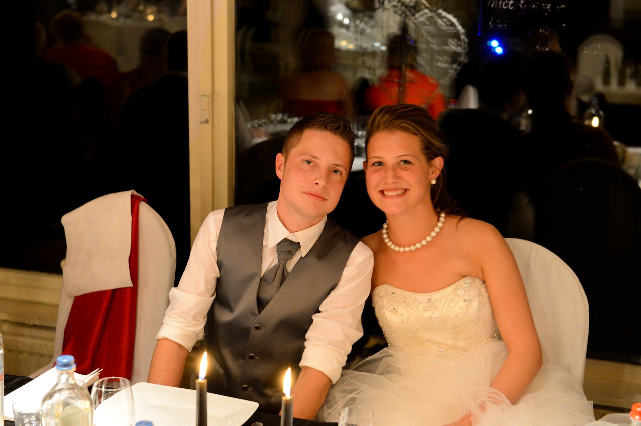 A newlywed couple sits together at their wedding reception, smiling and dressed in formal attire.