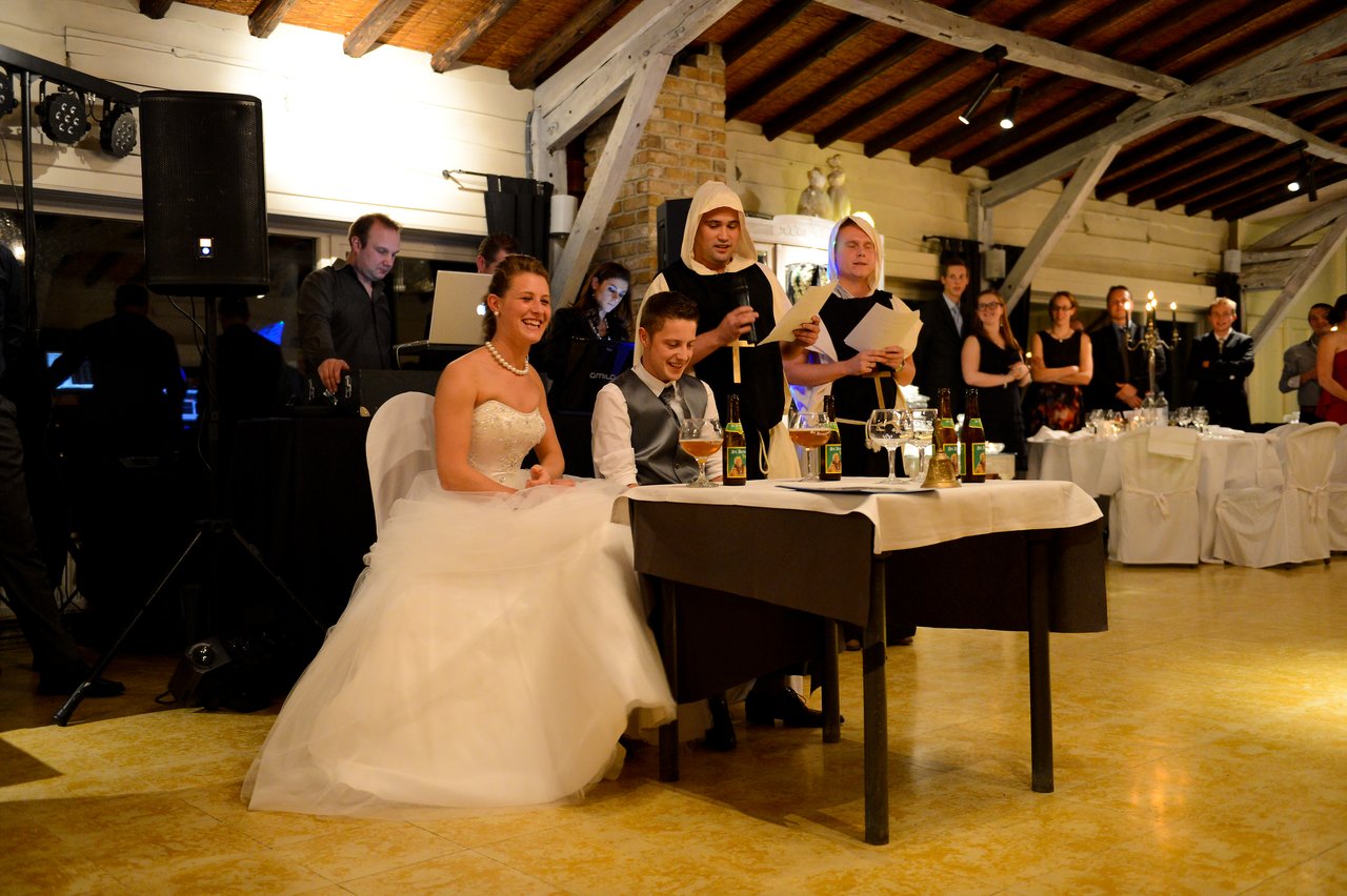 The bride and groom sit at a table, smiling, while two people in costumes read from papers at their wedding.