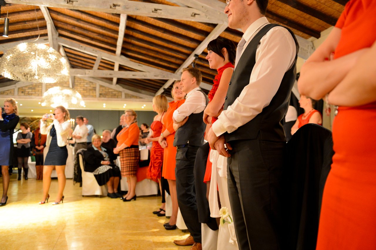 Wedding guests in formal attire stand and watch attentively, some smiling, during a celebration.