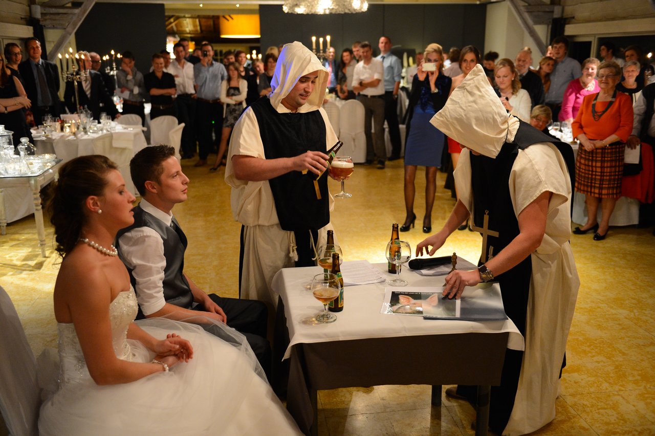 A bride and groom sit while two people in monk costumes perform a ceremony at a wedding reception.