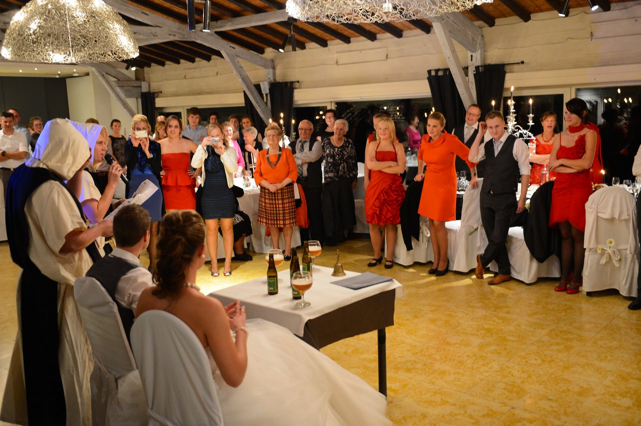 A woman in costume speaks into a microphone while the bride and groom listen, surrounded by wedding guests.