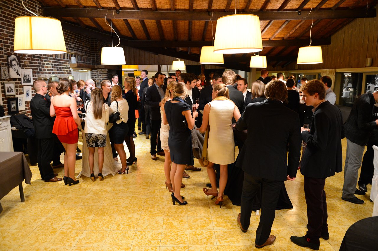 Guests in formal attire gather indoors, chatting and holding drinks at a wedding reception.