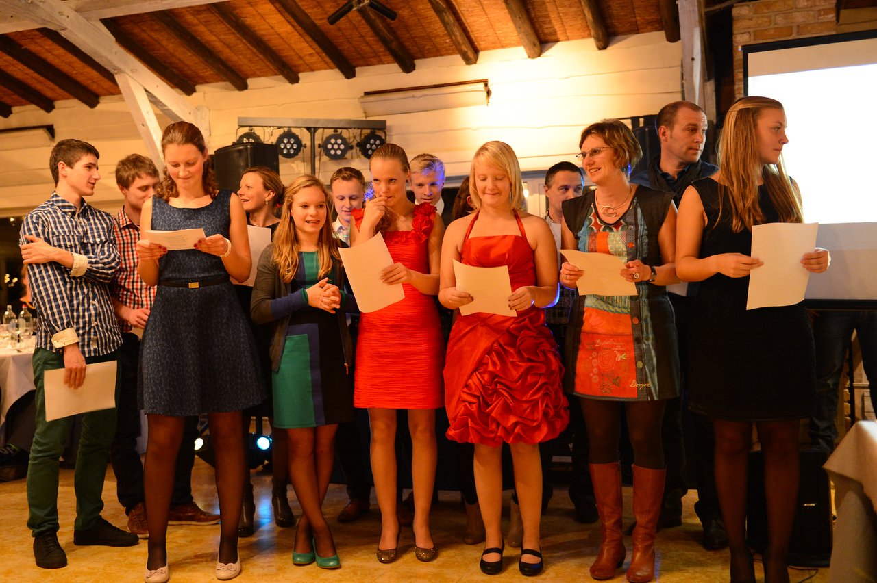 A group of people stand together, holding papers and reading or singing at a wedding celebration.