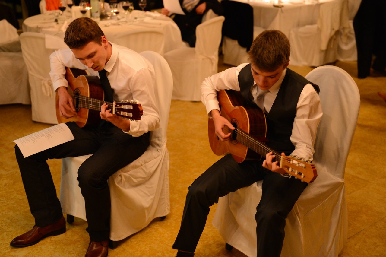 Two men in formal attire sit on chairs, playing acoustic guitars at a wedding reception.