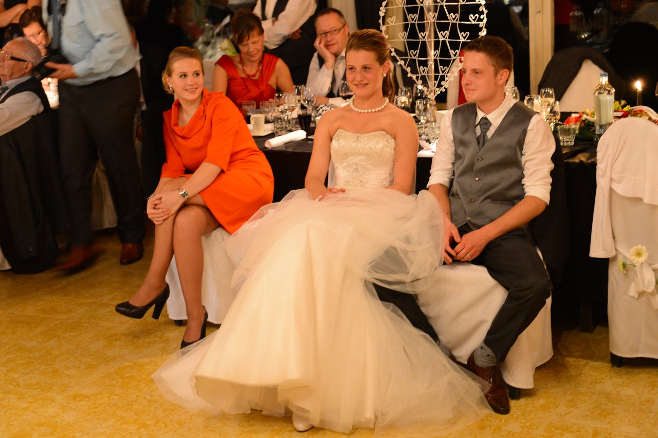 The bride and groom sit together at their wedding reception, smiling, with a guest in an orange dress beside them.