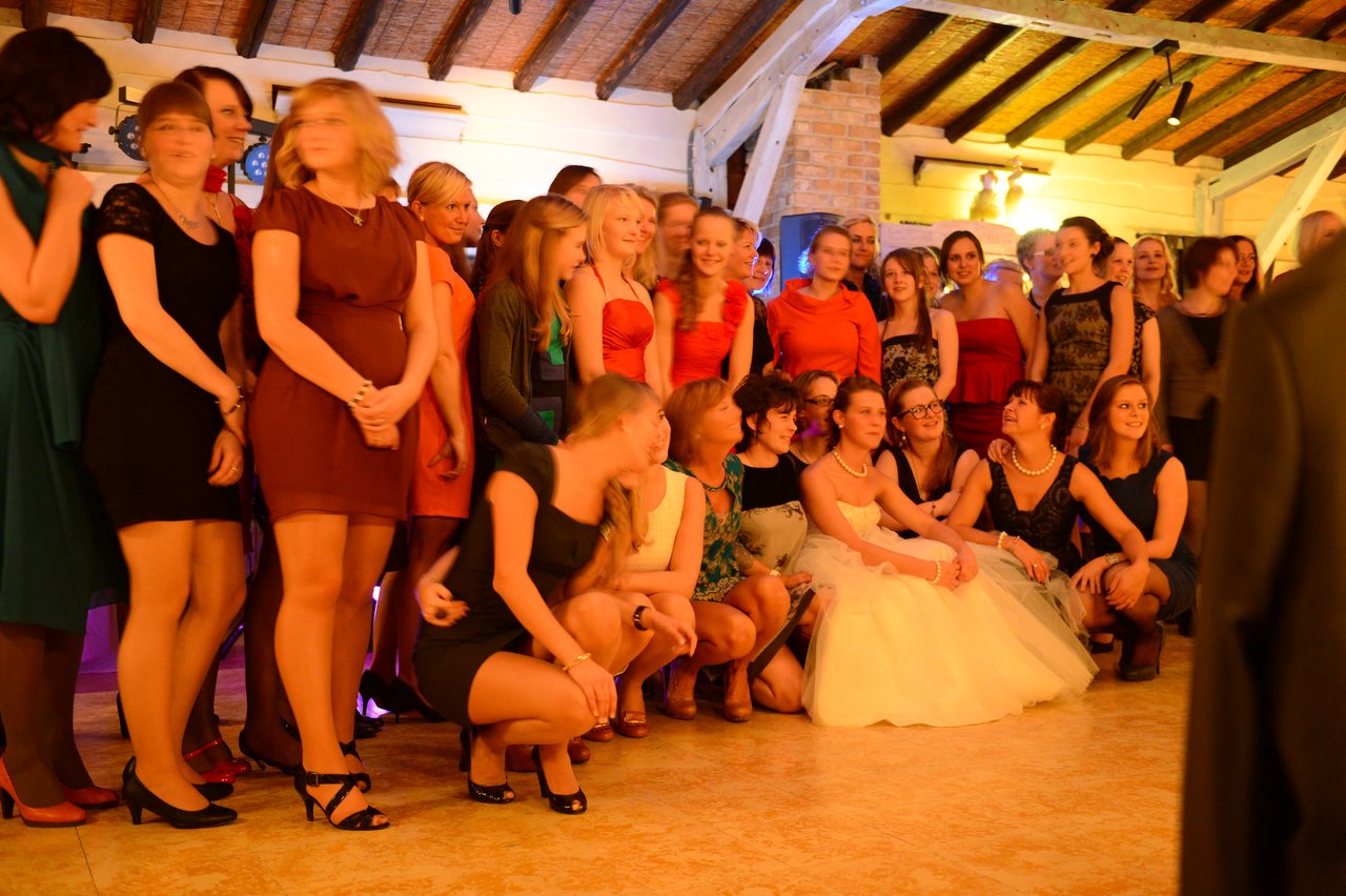 A bride in a white dress sits among a large group of women posing for a wedding photo indoors.
