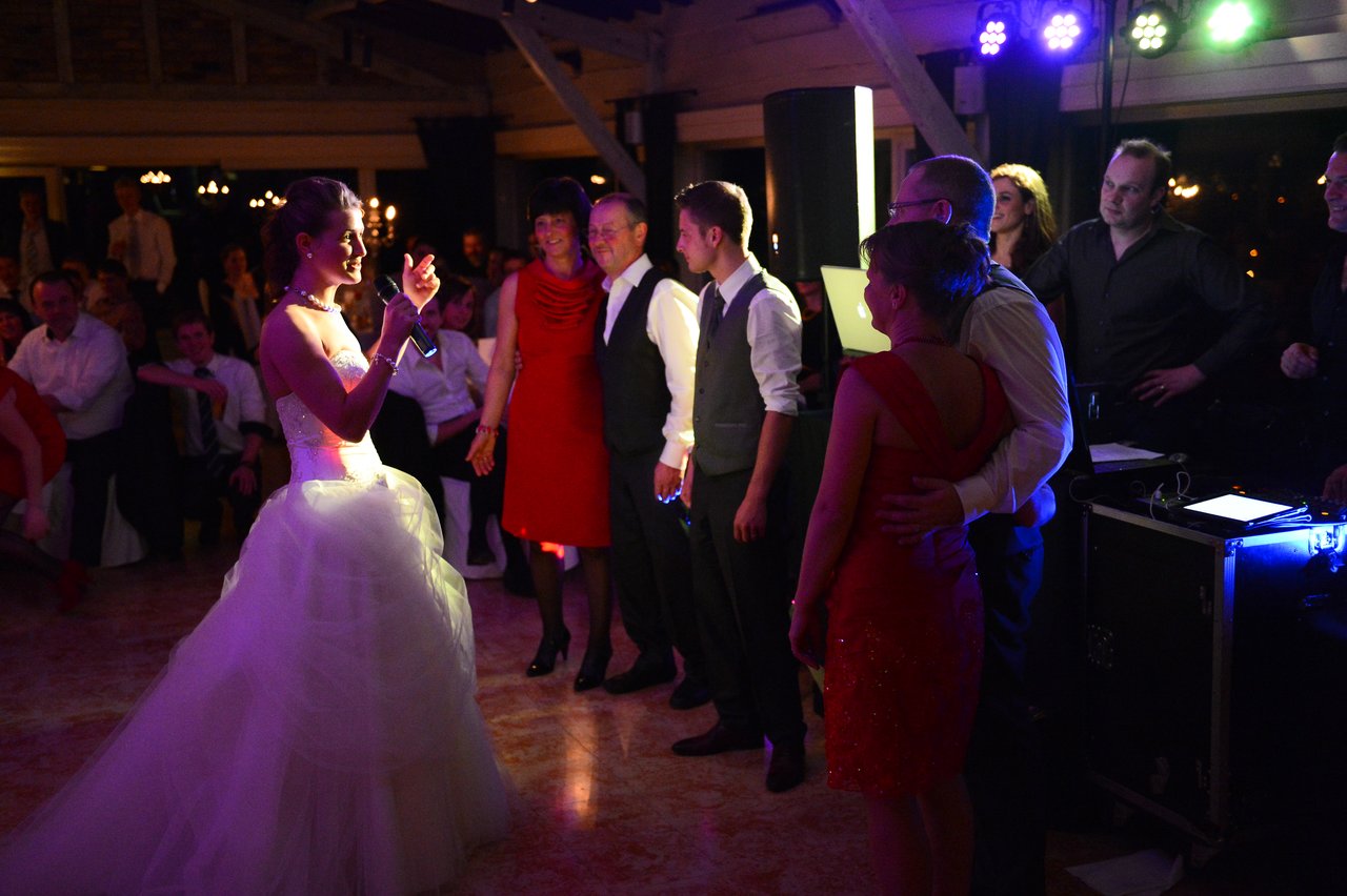 The bride in a white gown speaks into a microphone, addressing guests gathered on the dance floor.