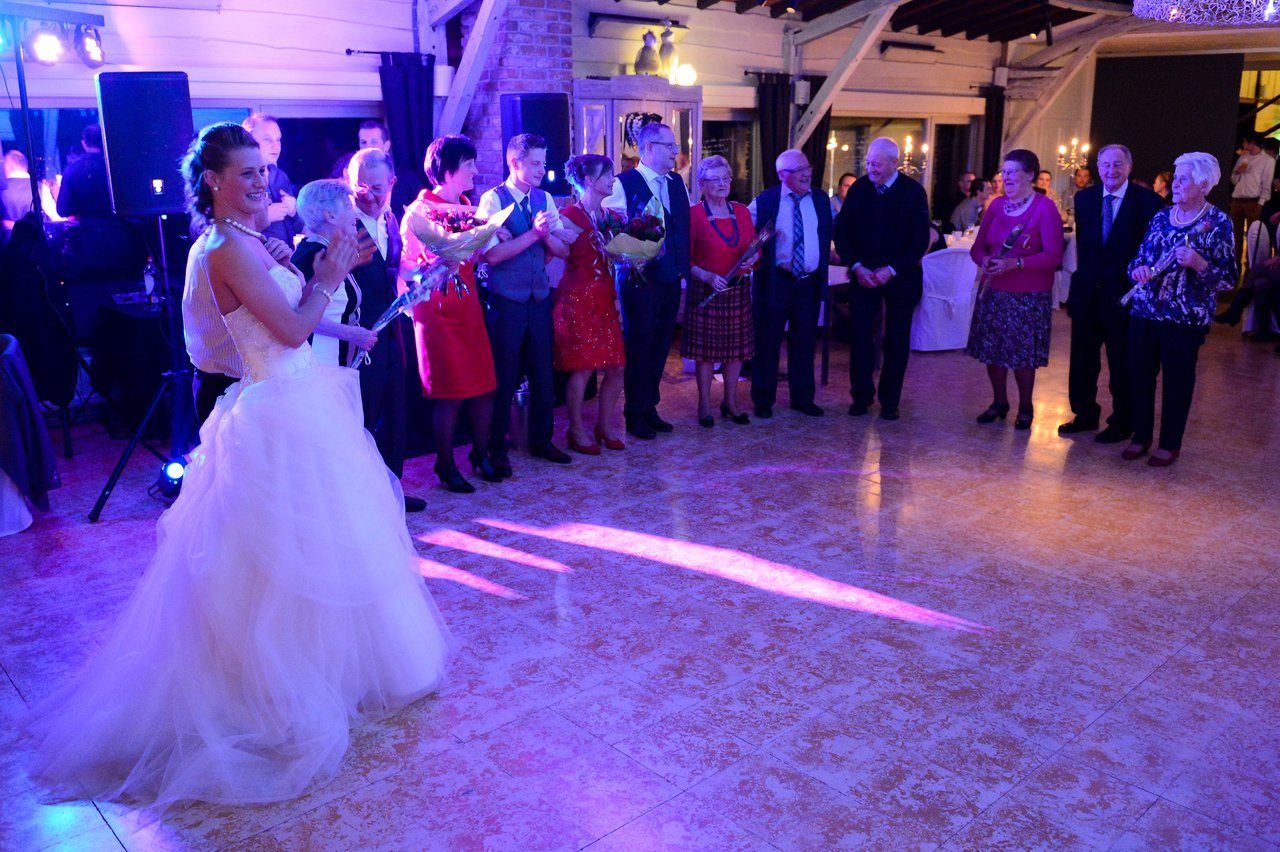 The bride in a white gown smiles and claps while guests stand in a line holding flowers at the wedding.