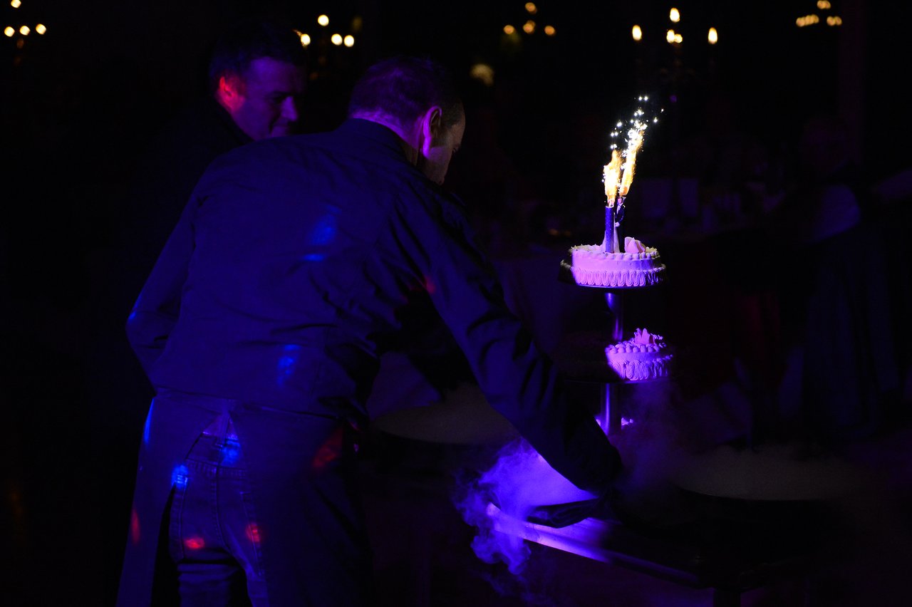 A waiter presents a tiered wedding cake with sparklers, surrounded by dramatic lighting and smoke effects.