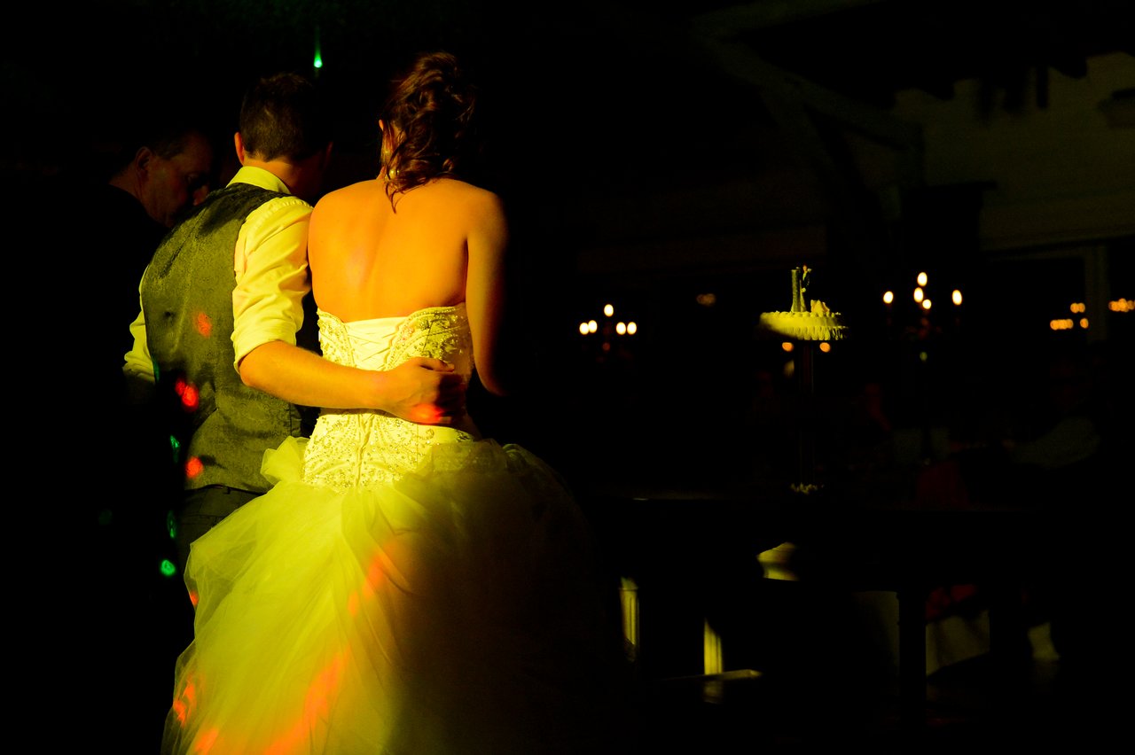 A newlywed couple stands close together, facing a wedding cake in a dimly lit reception venue.