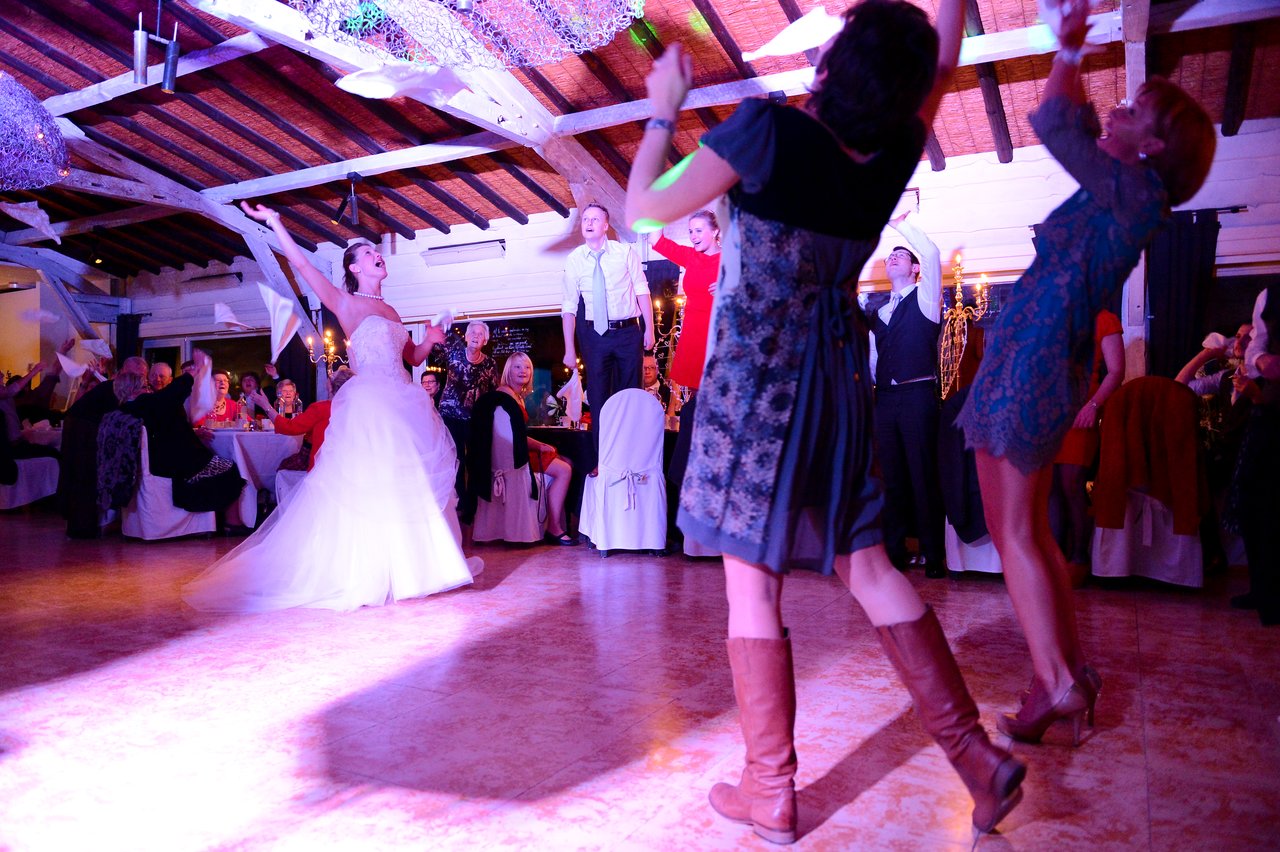 The bride in a white gown dances joyfully with guests, raising their hands and waving napkins in celebration.